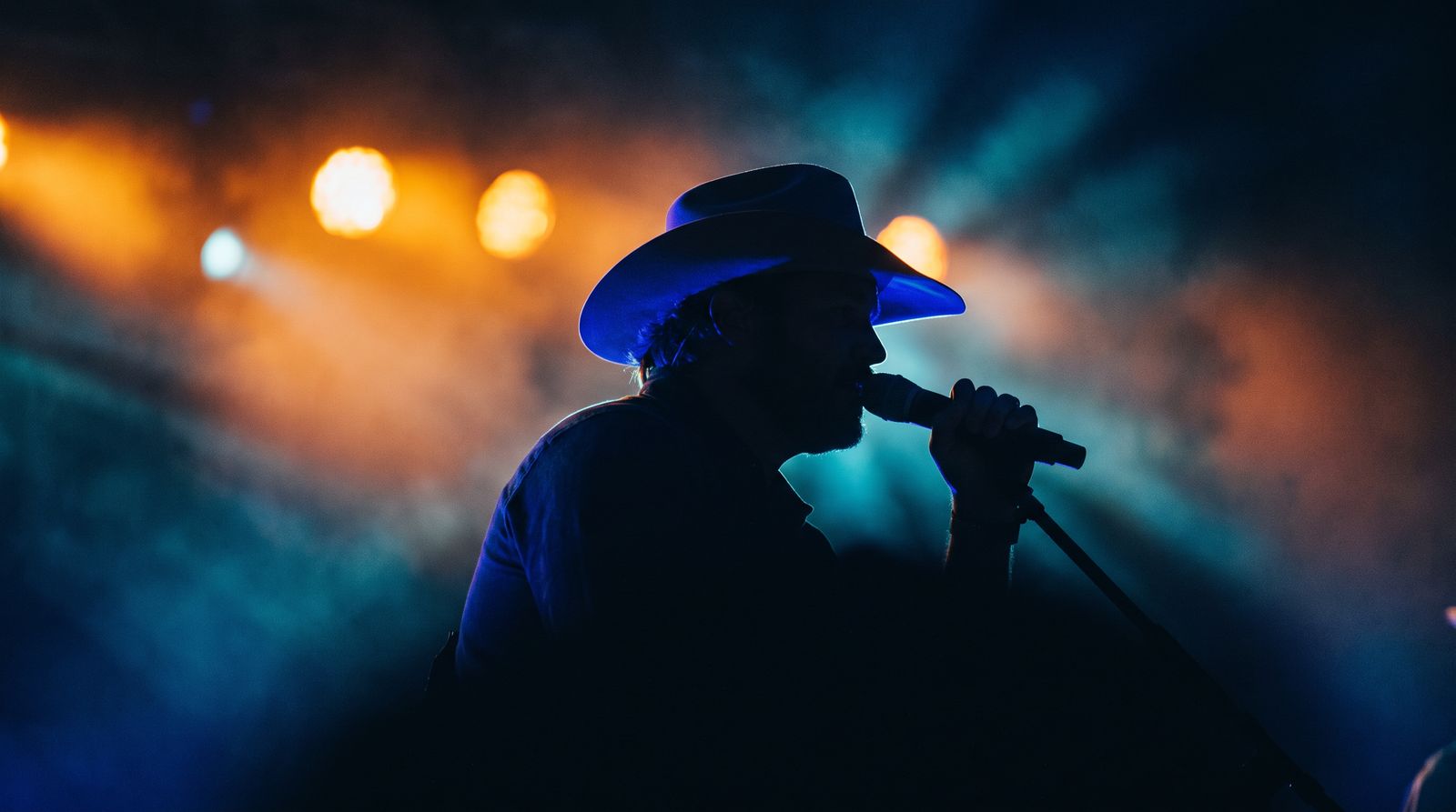 Cantante con sombrero vaquero de perfil y en silueta, cantando al micrófono frente a luces naranja desenfocadas.